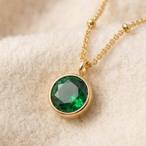 By Sophi Emerald Birthstone Pendant Necklace flat-lay on warm cream wool