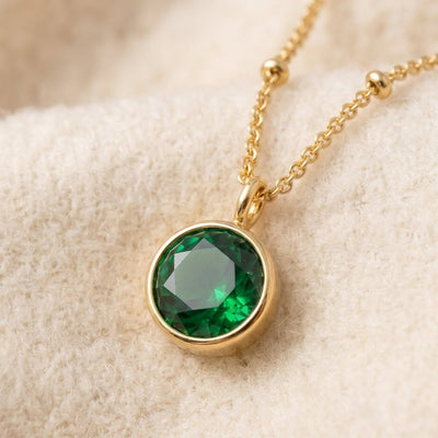 By Sophi Emerald Birthstone Pendant Necklace flat-lay on warm cream wool