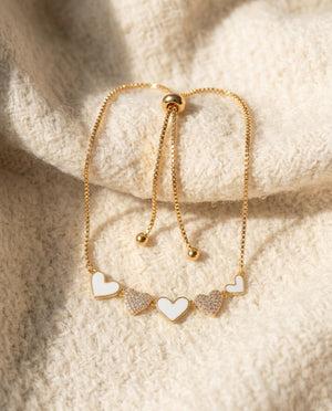By Sophi White Enamel & Gold Heart Bracelet flat-lay on warm cream wool